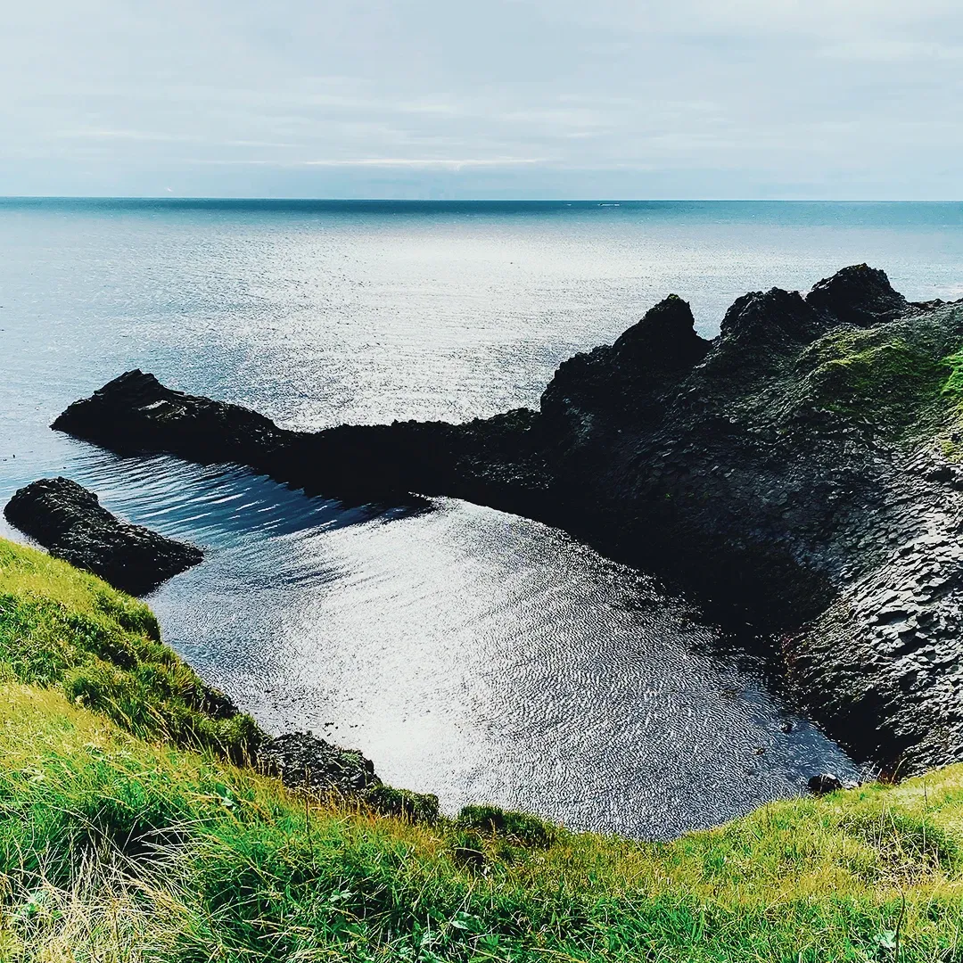 A small rocky coastal inlet with calm reflective water and green clifftops under a pale overcast sky