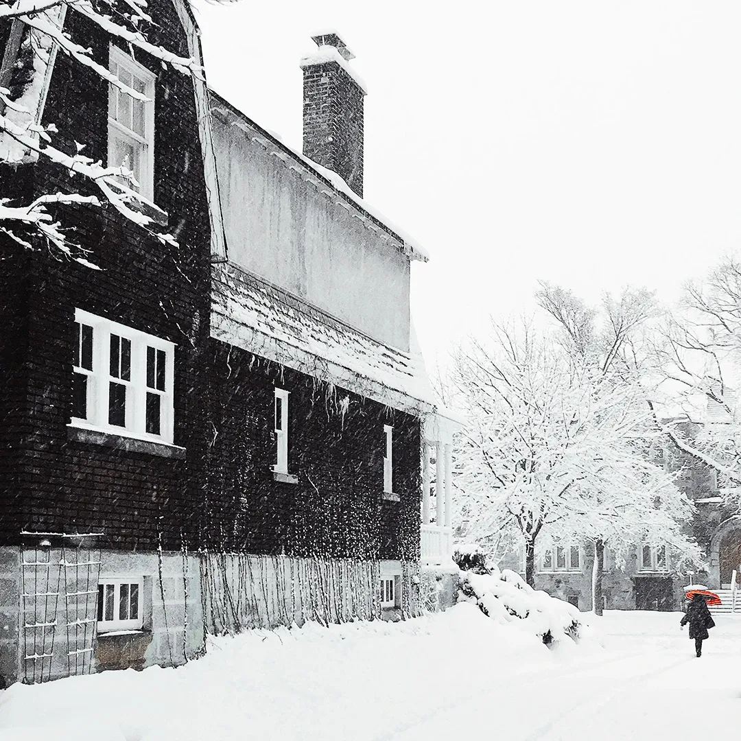 A lone figure with a red umbrella walks past a dark snow-blanketed house during a heavy winter snowfall