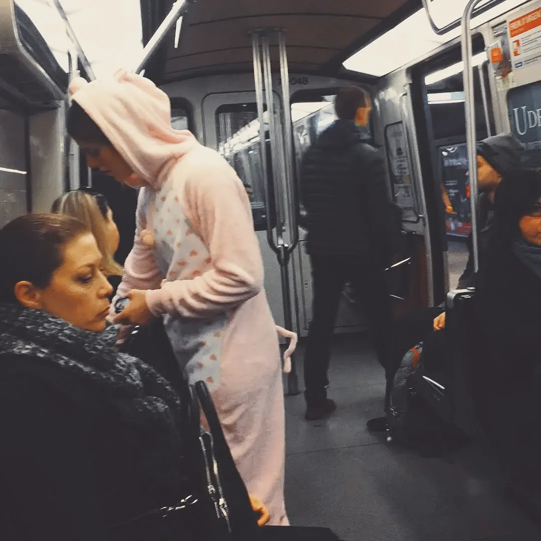 A person in a full pink unicorn onesie standing on a busy subway car surrounded by commuters in winter coats