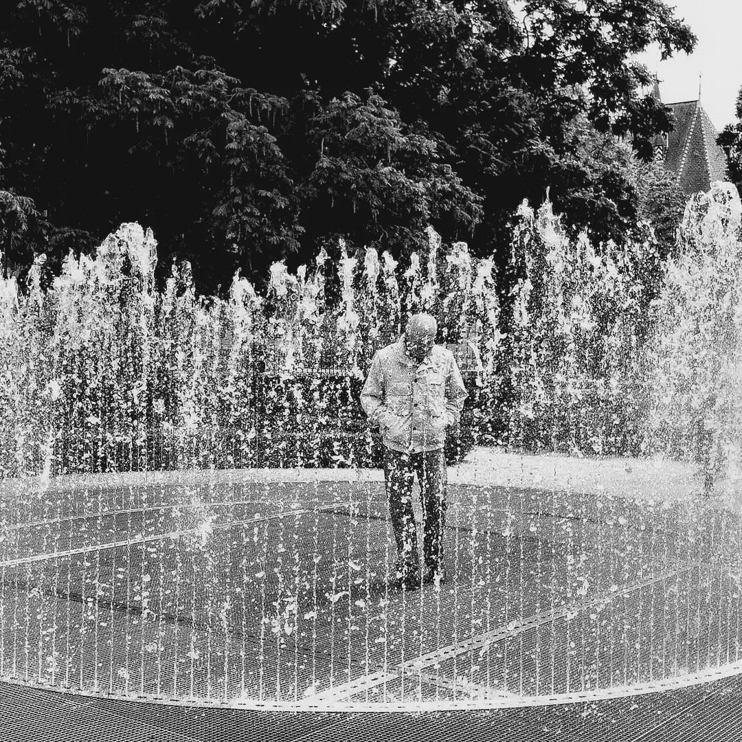 Black and white photo of an elderly man standing calmly inside a circular fountain with jets of water surrounding him