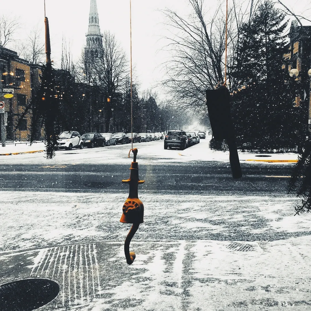 A small decorative figure hangs from a wire across a snowy Montreal street, a church steeple visible in the grey distance