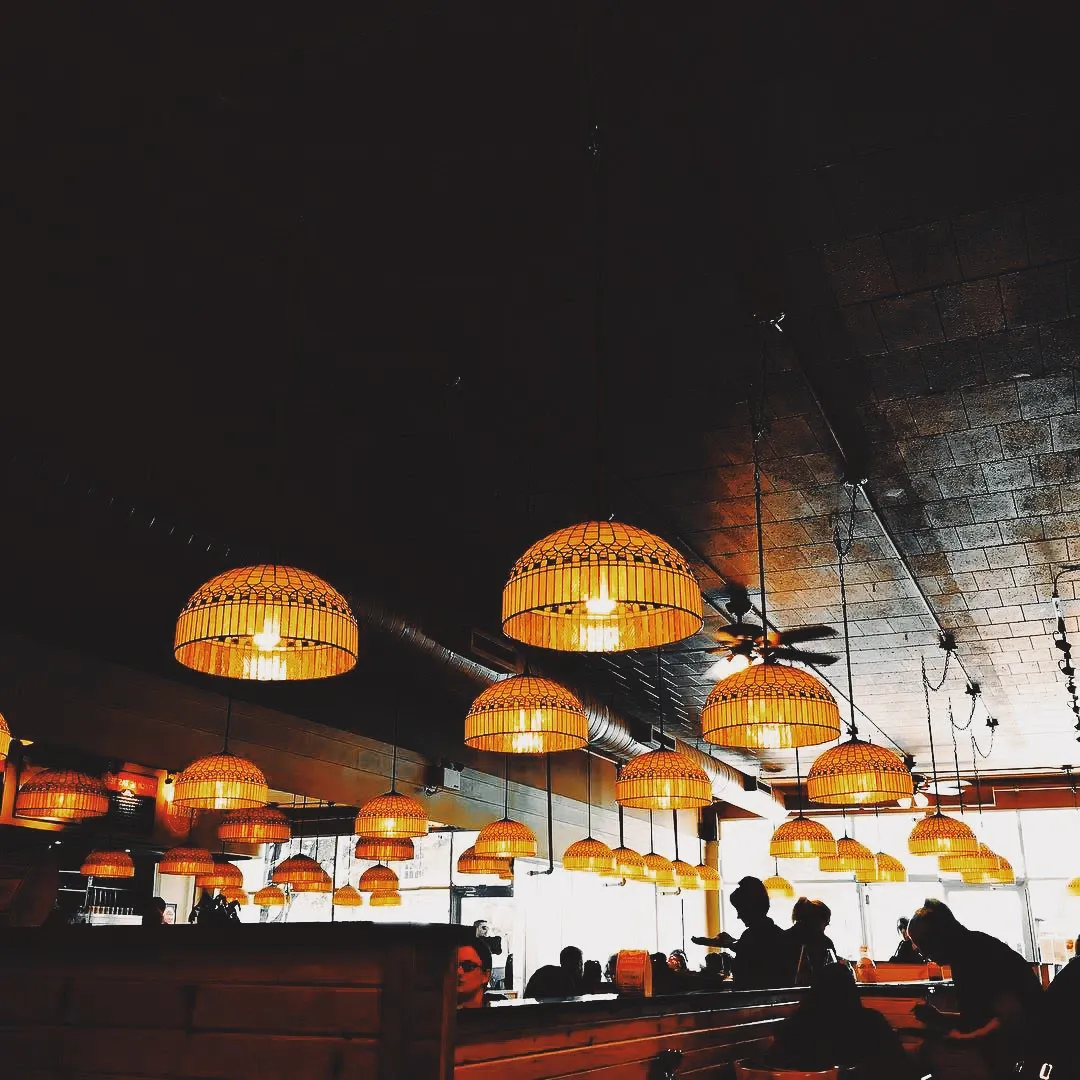 Rows of warm wicker pendant lamps hanging from a dark restaurant ceiling above the silhouettes of diners