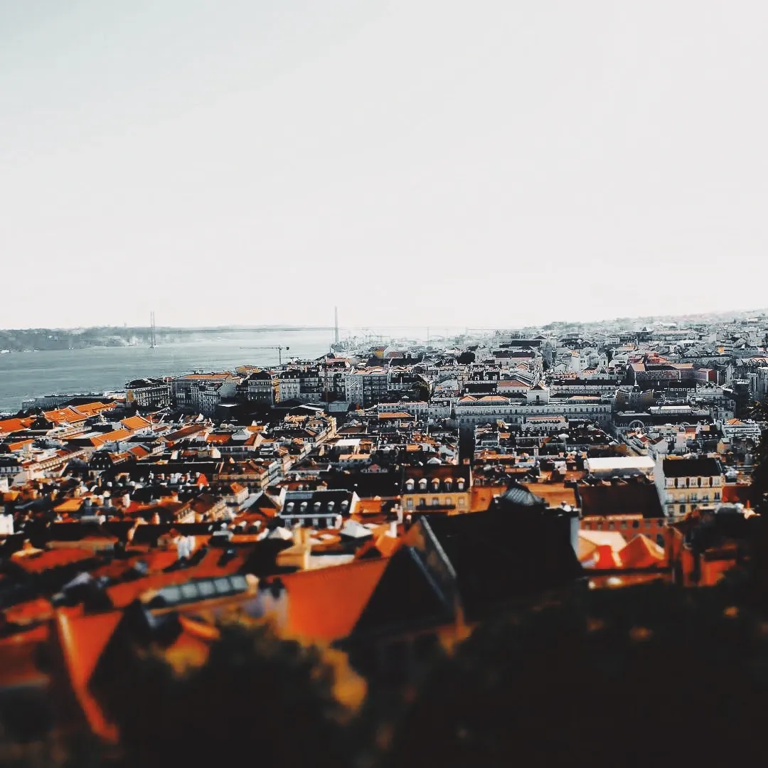 Tilt-shift aerial view of Lisbon's terracotta rooftops stretching toward the Tagus river and the 25 de Abril bridge