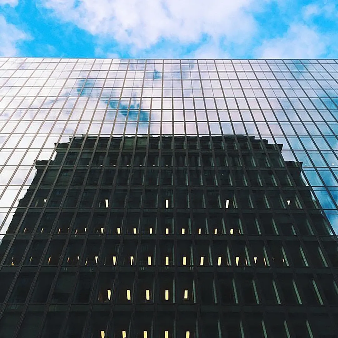 Glass-clad office tower reflecting a cloud-filled blue sky, with an older dark building visible through the facade