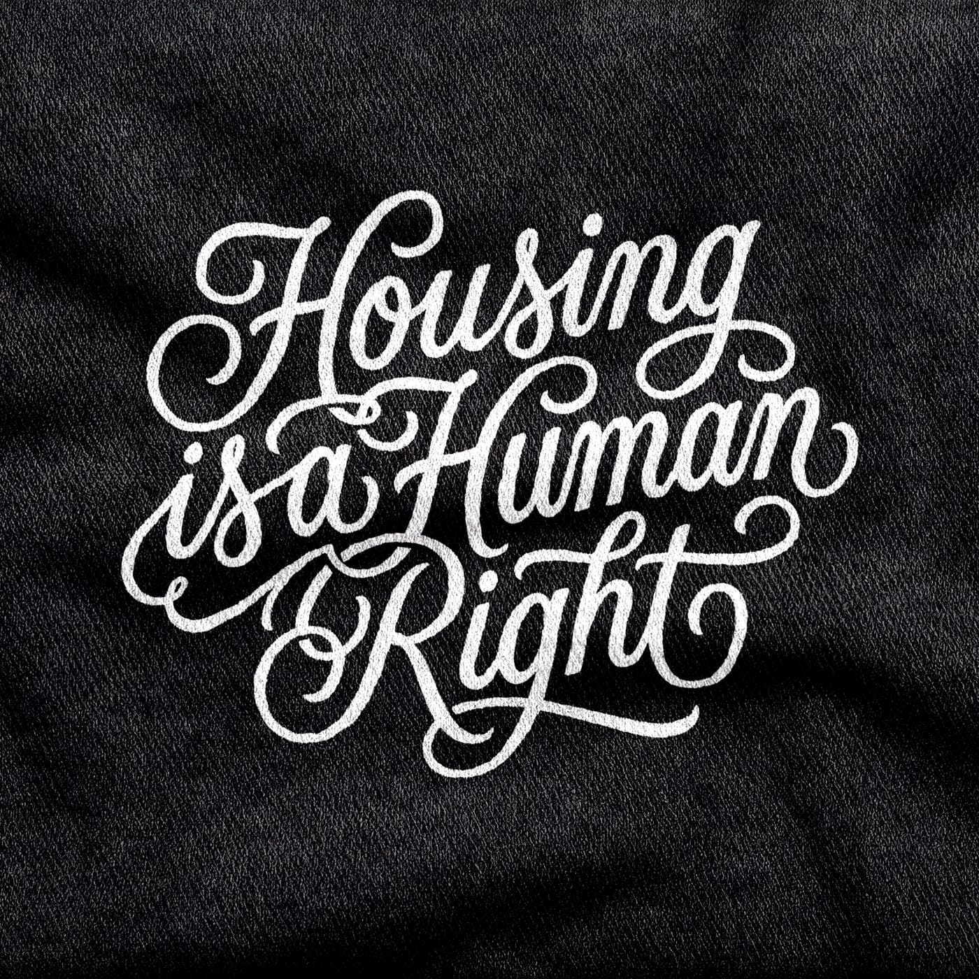 Ornate sign-painter's script reading "Housing is a Human Right", three lines deep and heavy with flourishes, white on a black tee.