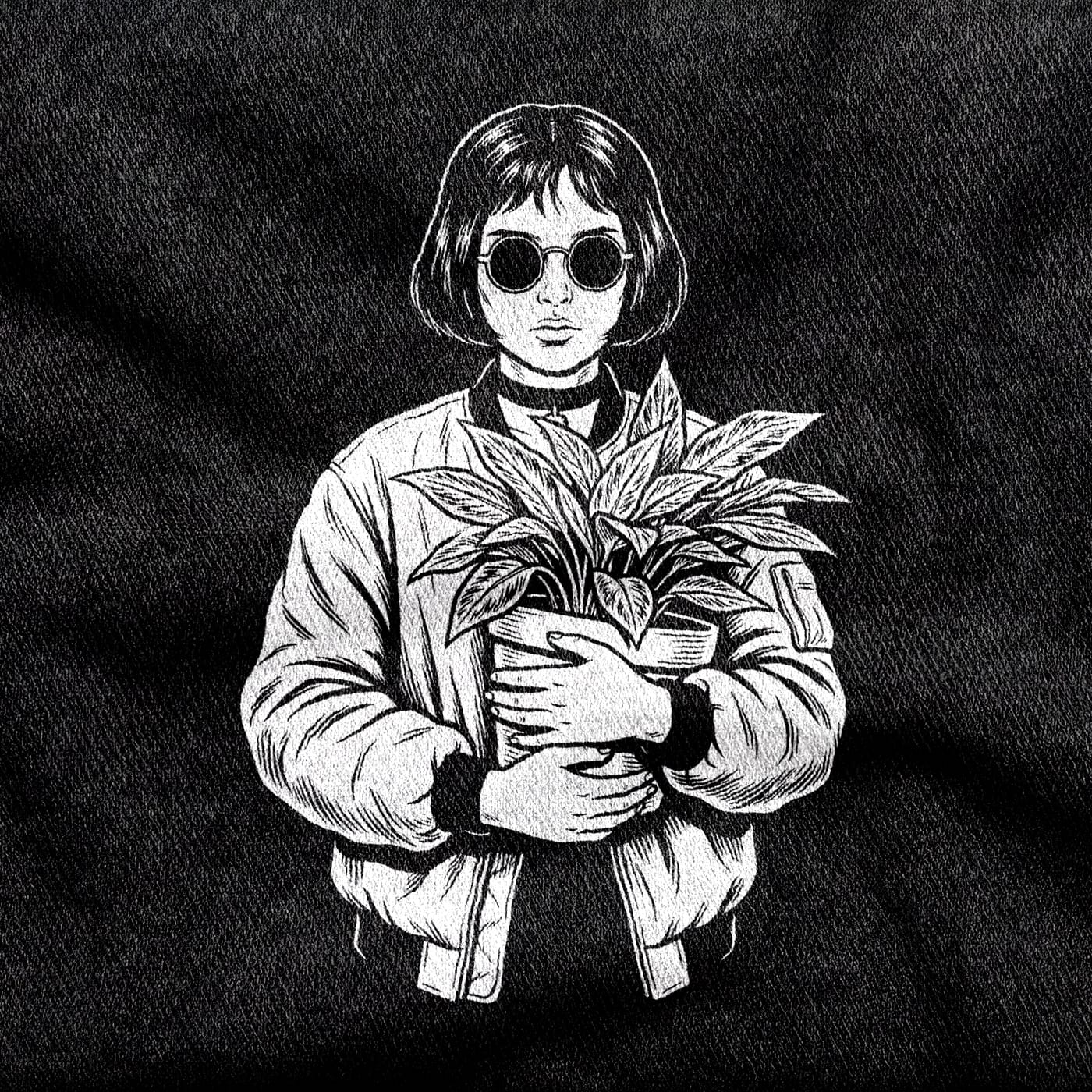 Mathilda with a blunt bob, round dark glasses, a choker and a bomber jacket, both arms wrapped around a potted plant, white on a black tee.