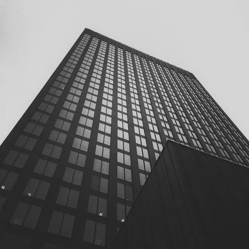 Black and white low-angle shot of a glass skyscraper's repeating window grid dissolving into a grey overcast sky