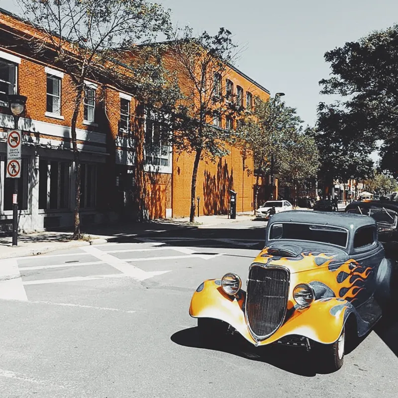 A vintage hot rod with yellow flame paint job parked on a sun-drenched urban street lined with brick buildings
