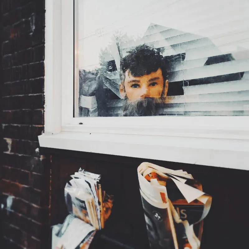 A bearded man peers curiously through a shop window, reflected in the glass alongside bundles of paper bags