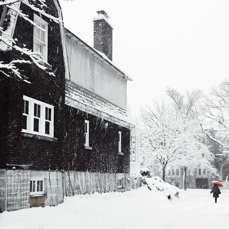 A lone figure with a red umbrella walks past a dark snow-blanketed house during a heavy winter snowfall