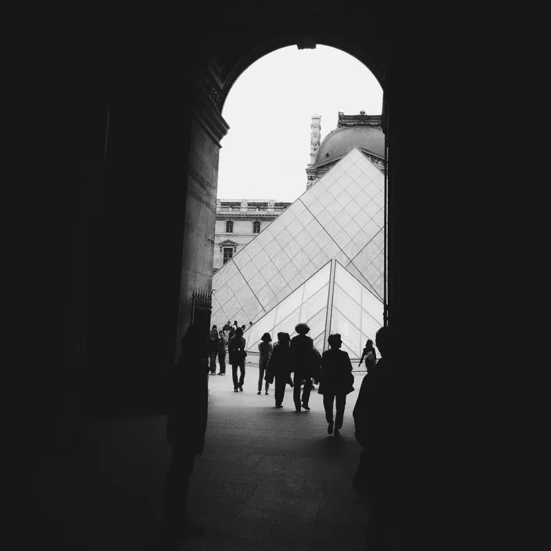 Black and white view of the Louvre Pyramid framed by a dark stone archway, silhouetted visitors crossing the courtyard