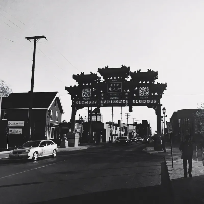 Black and white photo of a traditional Chinese gate spanning a street in Chinatown, a lone pedestrian walking beneath it