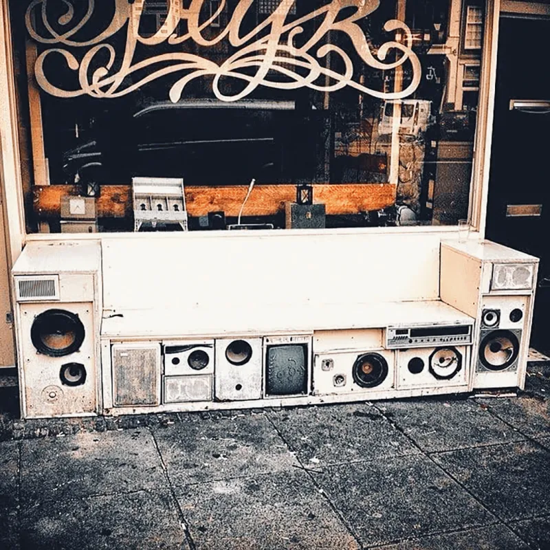 A bench made entirely of stacked vintage stereo speakers and radios sitting outside a shop with hand-lettered signage