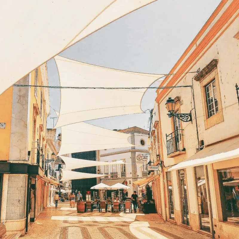 A sun-drenched pedestrian street in the Algarve covered by large shade sails, lined with cafes and colourful facades