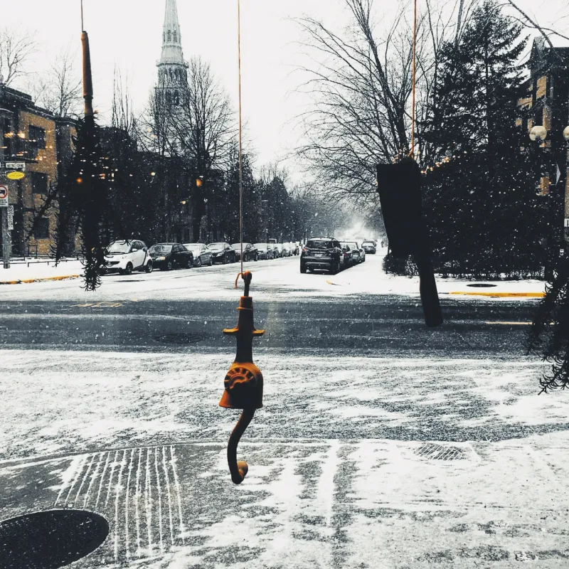 A small decorative figure hangs from a wire across a snowy Montreal street, a church steeple visible in the grey distance