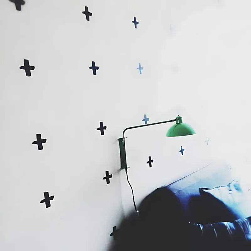 White bedroom wall covered in scattered black cross stickers beside a green swing-arm reading lamp