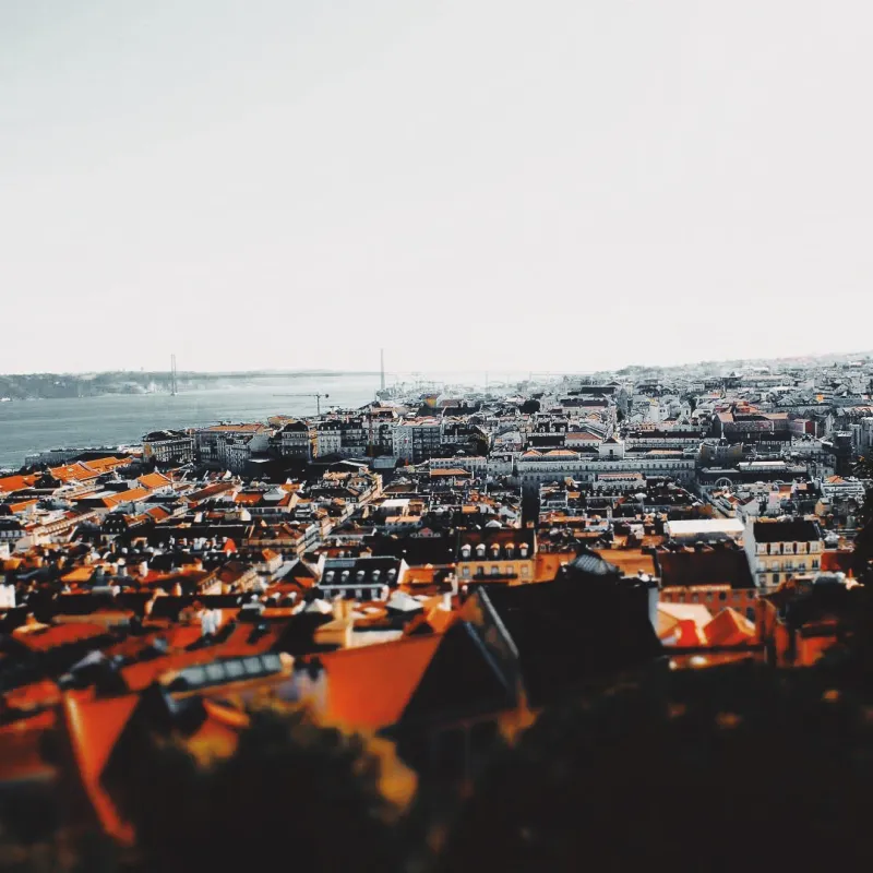 Tilt-shift aerial view of Lisbon's terracotta rooftops stretching toward the Tagus river and the 25 de Abril bridge