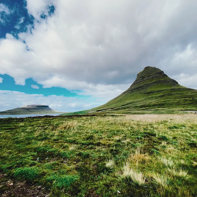 The iconic cone-shaped Kirkjufell mountain rising from green Icelandic grassland under a dramatic cloudy sky