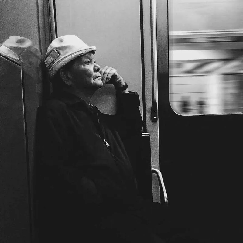 Black and white portrait of an older man in a flat cap leaning against a subway car door, lost in thought as the platform blurs past