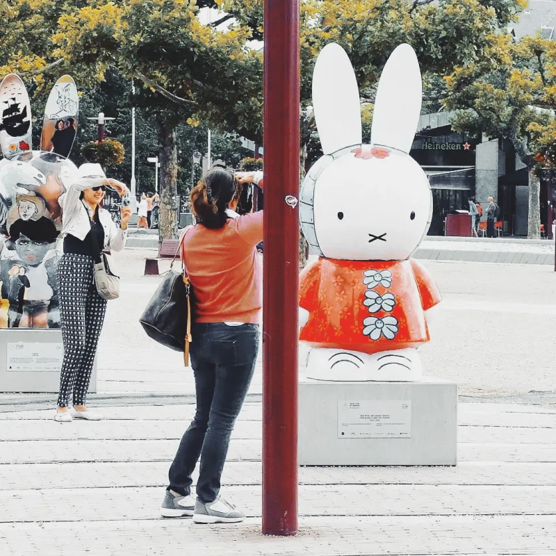 A woman photographing a large Miffy bunny sculpture in an orange floral dress on a sunny city square