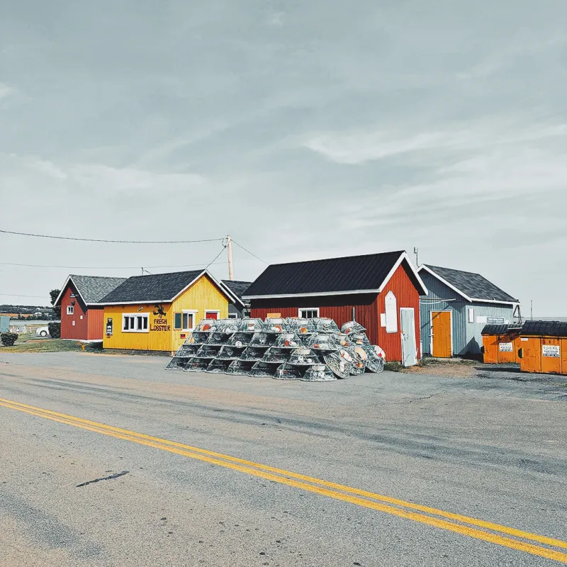 Colourful wooden fishing sheds and stacked lobster traps along a quiet coastal road under an overcast sky