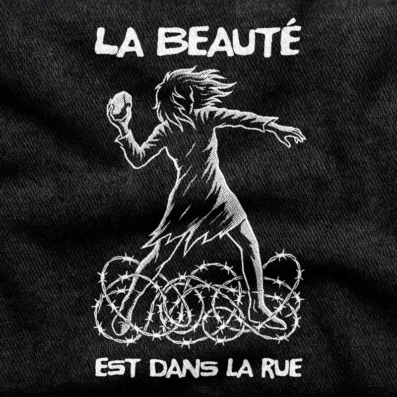 A woman mid-throw with storming hair above barbed wire, the May 68 slogan "La Beauté est dans la Rue", white on a black tee.