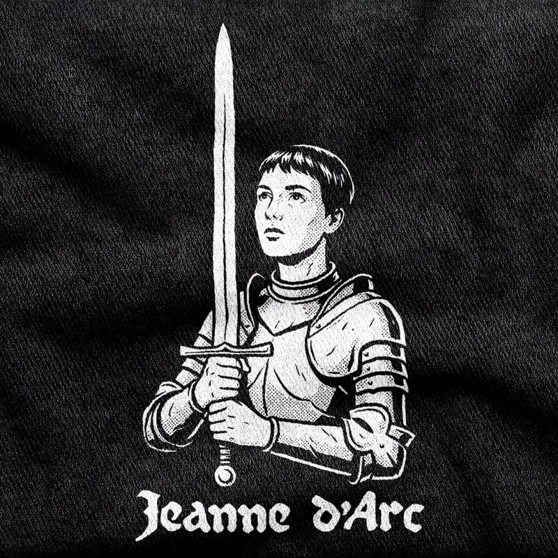 Joan of Arc in plate armour with cropped hair, her sword held upright like a cross, under a blackletter wordmark, white on a black tee.