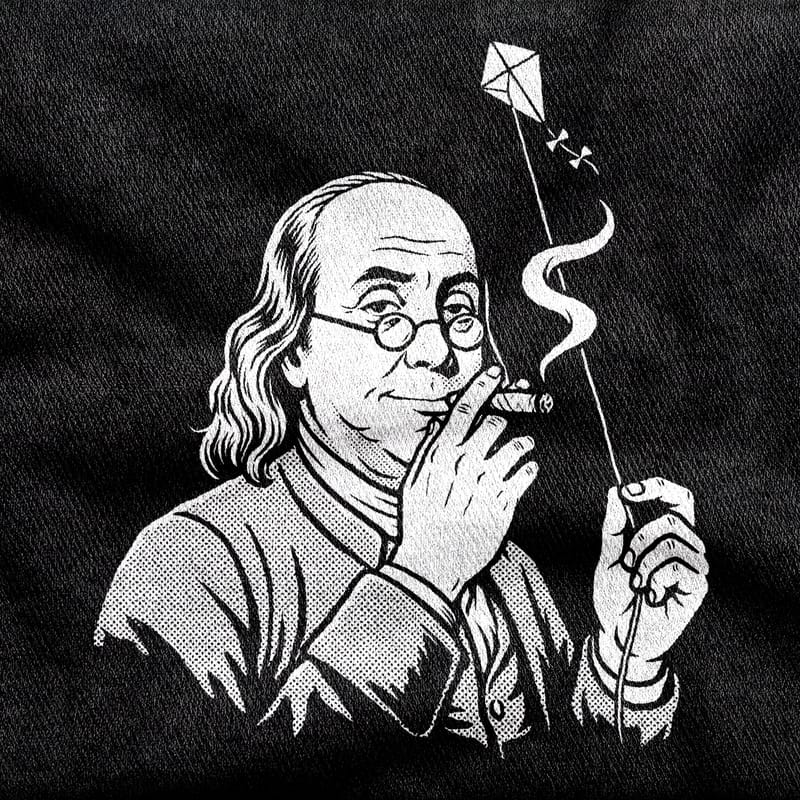 Benjamin Franklin in spectacles and a colonial coat, a kite string in one hand and something rolled and lit in the other, white on a black tee.