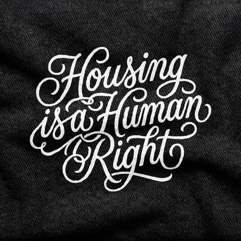 Ornate sign-painter's script reading "Housing is a Human Right", three lines deep and heavy with flourishes, white on a black tee.