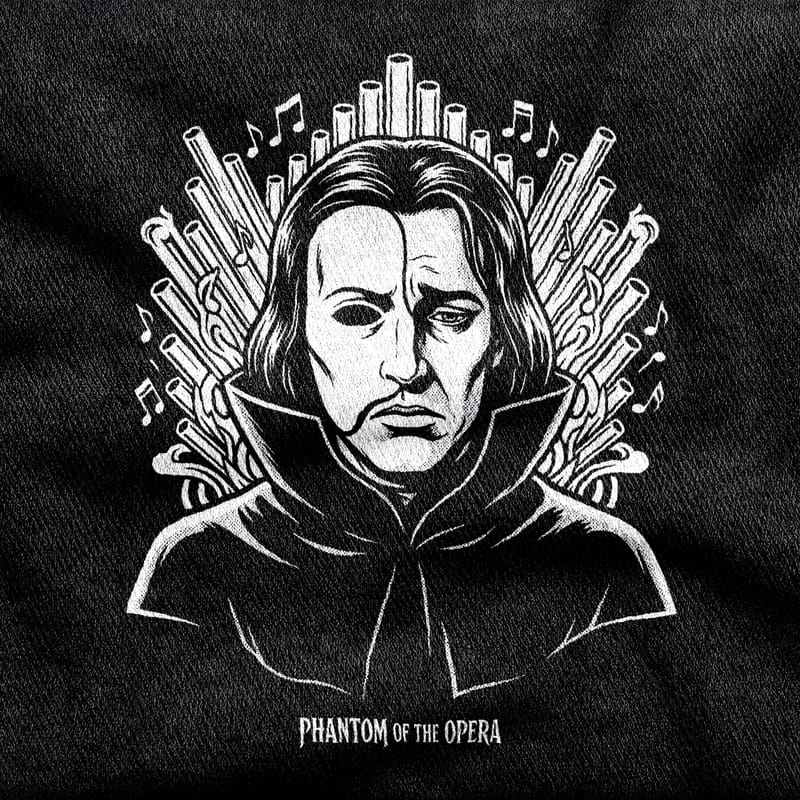 The Phantom of the Opera in a cape with half his face in shadow, backed by a fan of organ pipes and drifting notes, white on a black tee.