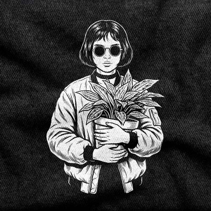 Mathilda with a blunt bob, round dark glasses, a choker and a bomber jacket, both arms wrapped around a potted plant, white on a black tee.